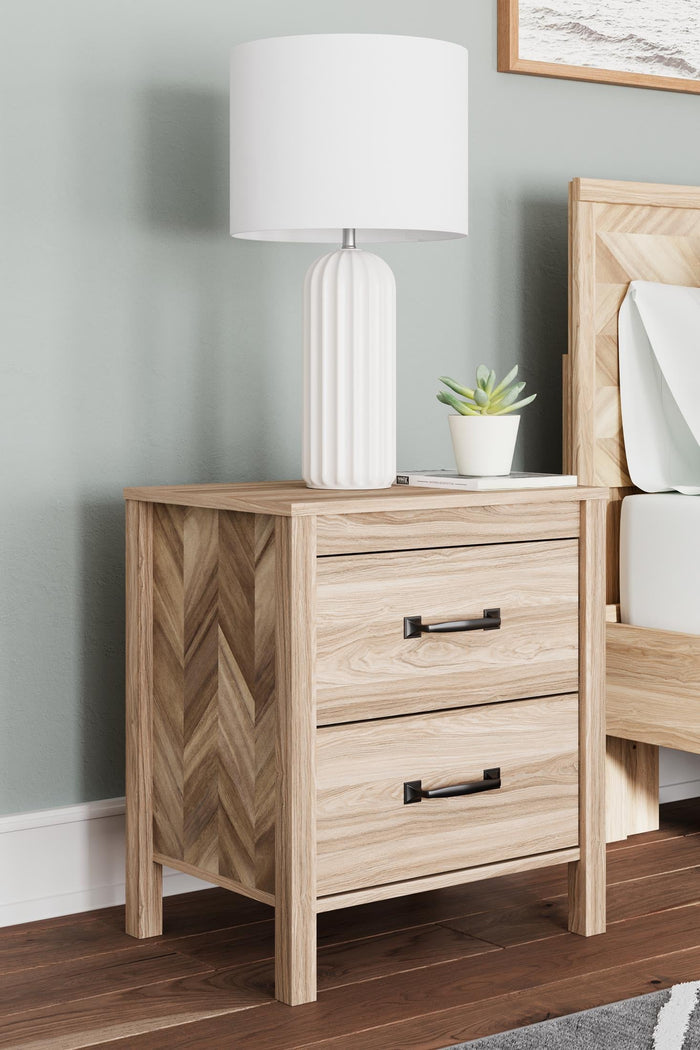 Light wood nightstand with two drawers and black bar pulls, with lamp on top