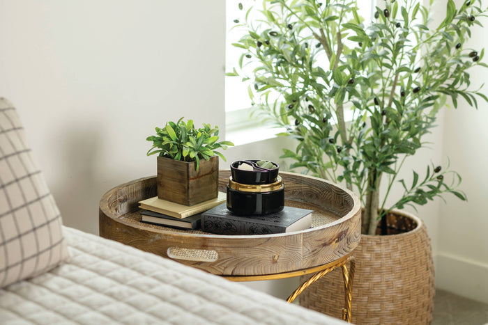 A round wooden side table with a gold metal frame, styled in a bright room next to a bed with a plaid pillow and a large potted olive tree.