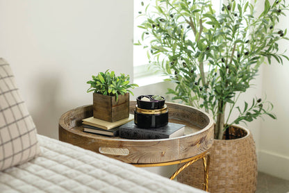 A round wooden side table with a gold metal frame, styled in a bright room next to a bed with a plaid pillow and a large potted olive tree.