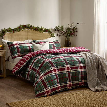A bed dressed in a festive plaid comforter set with red, green, white, and dark blue tartan patterns, styled in a cozy bedroom with a woven cane headboard, holiday greenery garland, and a gray throw blanket.