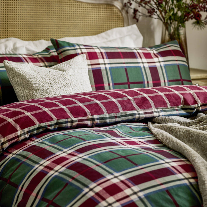 A styled bed with plaid bedding in deep red, green, cream, and blue tartan patterns, including a textured cream pillow, a woven cane headboard, and a soft gray throw blanket.