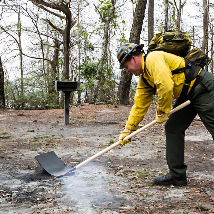 Wildland firefighter using fire swatter tool to suppress residual ground heat in a forested area.