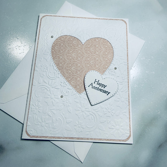 An embossed white anniversary card with a large beige patterned heart and a smaller white heart reading 'Happy Anniversary' on a light gray surface, with white envelopes behind it.