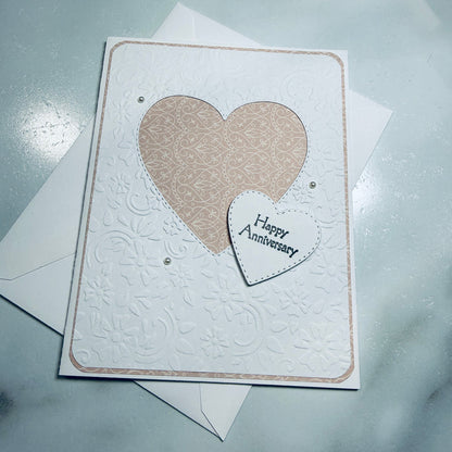 An embossed white anniversary card with a large beige patterned heart and a smaller white heart reading 'Happy Anniversary' on a light gray surface, with white envelopes behind it.