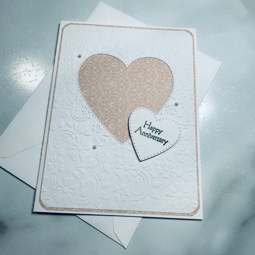 An embossed white anniversary card with a large beige patterned heart and a smaller white heart reading 'Happy Anniversary' on a light gray surface, with white envelopes behind it.