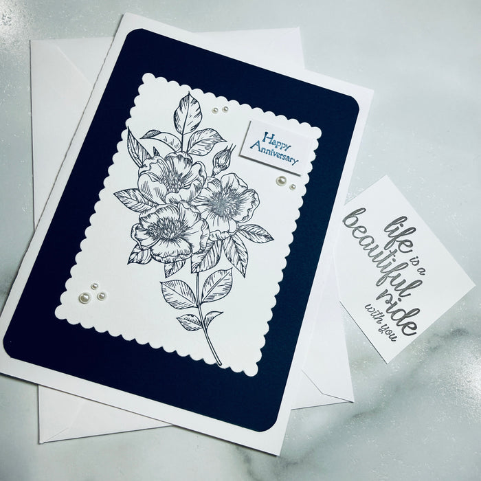 A handmade anniversary greeting card with a navy blue and white layered design, featuring a black floral line drawing and the words 'Happy Anniversary' on the main card, shown with a smaller card reading "Life is a beautiful ride with you" and envelopes on a light marble surface.
