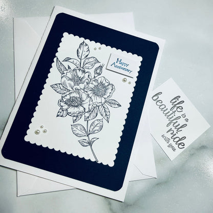 A handmade anniversary greeting card with a navy blue and white layered design, featuring a black floral line drawing and the words 'Happy Anniversary' on the main card, shown with a smaller card reading "Life is a beautiful ride with you" and envelopes on a light marble surface.