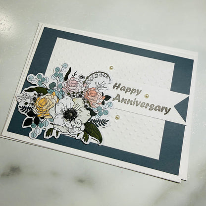 A handmade square anniversary card with a navy blue border, white center panel, and layered floral cutouts in white, pink, yellow, and green. The card reads 'Happy Anniversary' in metallic lettering and is photographed on a light marble-like surface.