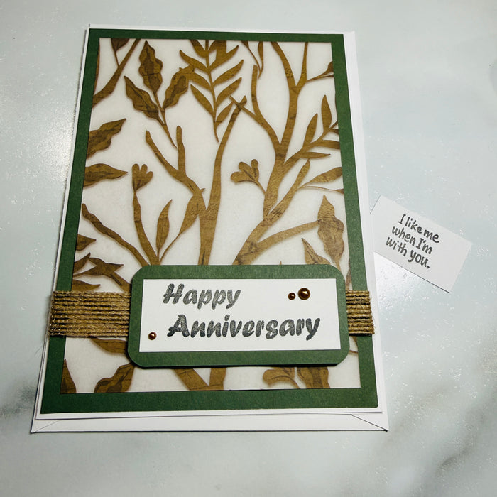 An anniversary greeting card with a laser-cut tree design in olive green and gold, featuring a central label that reads 'Happy Anniversary' and a small card beside it reading "i like me when im with you" on a light marble surface.