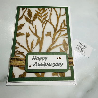An anniversary greeting card with a laser-cut tree design in olive green and gold, featuring a central label that reads 'Happy Anniversary' and a small card beside it reading "i like me when im with you" on a light marble surface.