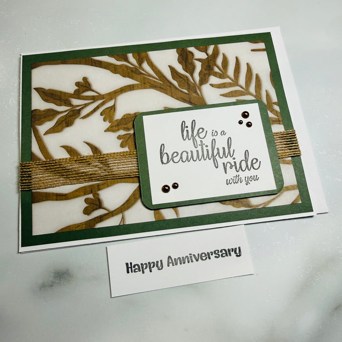 A handmade greeting card with a white base, dark green border, and laser-cut botanical leaf pattern in olive green. A small layered white label on the front reads 'life is a beautiful ride with you' in gray script, accented with tiny black dots and wrapped with twine.