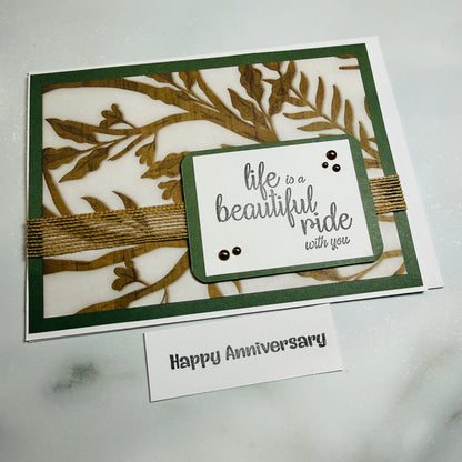 A handmade greeting card with a white base, dark green border, and laser-cut botanical leaf pattern in olive green. A small layered white label on the front reads 'life is a beautiful ride with you' in gray script, accented with tiny black dots and wrapped with twine.