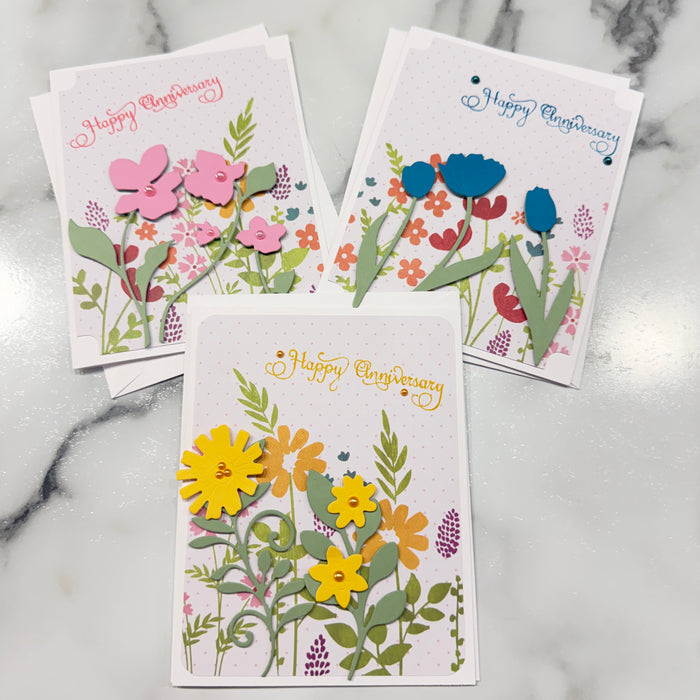 Three layered paper greeting cards with colorful floral designs and 'Happy anniversary' text, arranged on a white marble surface.