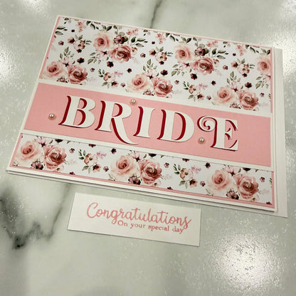 A handmade congratulations card featuring a floral rose pattern in pink and white, with the word 'BRIDE' cut out in the center and a separate matching 'Congratulations' card beside it, displayed on a light marble surface.
