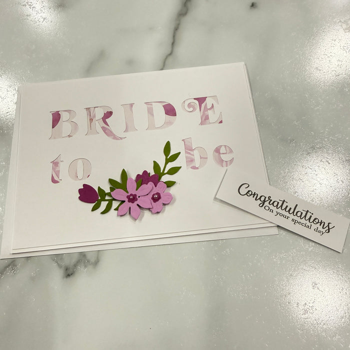 A white card with the words 'BRIDE to be' in a layered paper-cut style design, decorated with pink and purple flowers and green leaves, placed on a light marble surface next to a small white 'Congratulations' card.