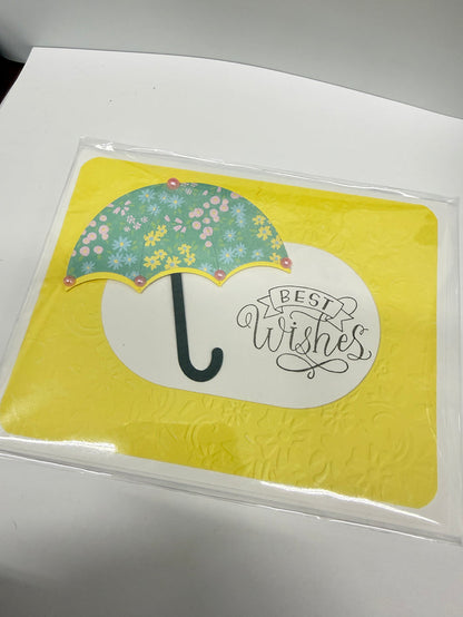 A square yellow card or coaster inside a clear plastic sleeve, featuring a white oval with the phrase 'BEST WISHES' in script and a floral green umbrella illustration on a white surface.
