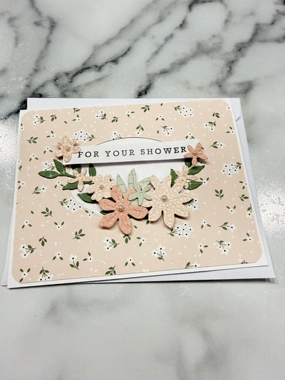 A handmade floral shower card with layered paper flowers and green leaves on a beige patterned background, placed on a white marble surface.