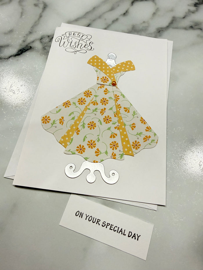 A handmade greeting card featuring a stylized dress illustration in a yellow and orange floral pattern on a white card, placed on a gray marble surface next to a small white card that reads 'ON YOUR SPECIAL DAY'.