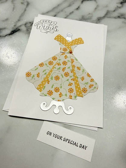 A handmade greeting card featuring a stylized dress illustration in a yellow and orange floral pattern on a white card, placed on a gray marble surface next to a small white card that reads 'ON YOUR SPECIAL DAY'.