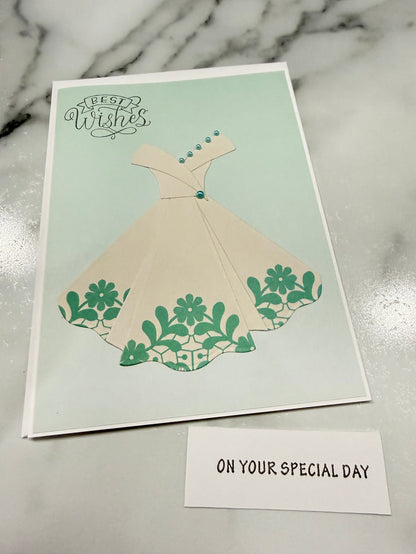 A square greeting card featuring a cream-colored dress with green floral patterns on a pale mint background, placed on a white marble surface next to a small white card that reads 'ON YOUR SPECIAL DAY'.