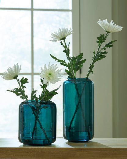 Two teal glass vases with white daisy stems on a wooden tabletop