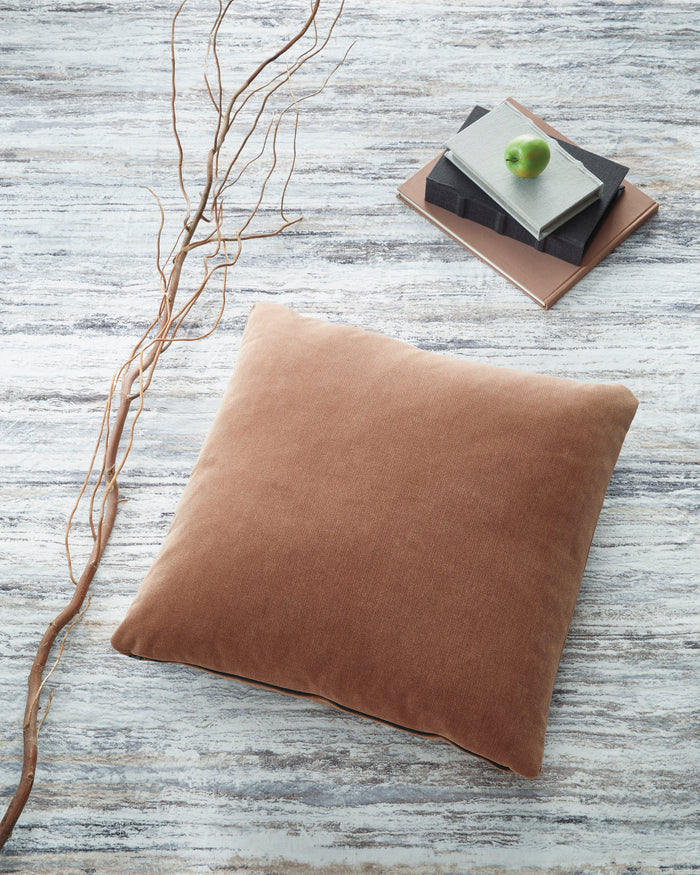 Square throw pillow in camel tan fabric, shown lying on a light blue surface.