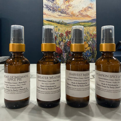 Four amber glass spray bottles with white labels and gold collars arranged on a white surface, with a colorful landscape painting hanging on the wall behind them.