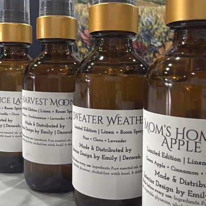 Four amber glass spray bottles with black atomizers and gold collars, lined up on a marble surface in front of a colorful landscape painting. The front bottle has a white label reading 'MOM'S HOME APPLE'.