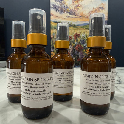 Five amber glass spray bottles with white labels and gold collars arranged on a light marble surface, with a dark wall and colorful landscape painting in the background.