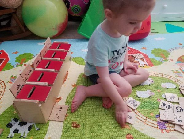 Toddler sitting on a colorful play mat, reaching for wooden cards next to a sorting box with red slots.