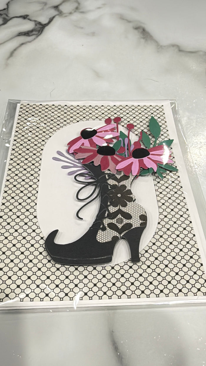 A decorative art print or card featuring a black high-heeled shoe filled with pink flowers and green leaves, placed on a patterned black-and-white background, photographed on a marble surface.
