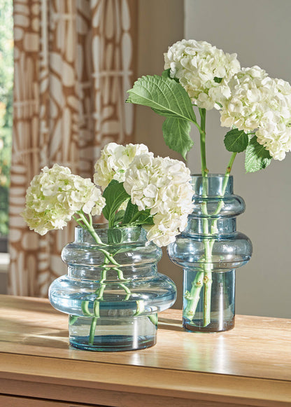 Blue glass vase with two stems of white hydrangea-style flowers and green leaves