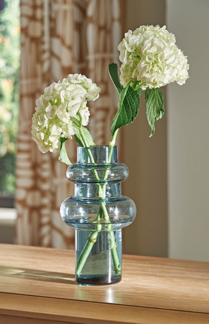 Blue glass vase with white hydrangea-style flowers and green leaves, ribbed rings